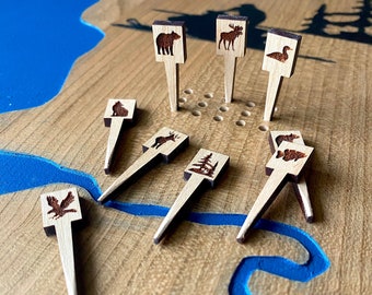Può includere: Pedine di gioco in legno con disegni di animali e alberi, tra cui un orso, un alce, un'anatra e un'aquila. I pezzi sono posizionati su una tavola di legno con accenti blu. Il tabellone di gioco ha il testo "Lakes Cabin".