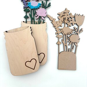 May include: Two wooden mason jar shapes with a heart design. One jar is layered on top of the other. A separate wooden cutout of a bouquet of flowers with pink, purple, blue, and yellow flowers.