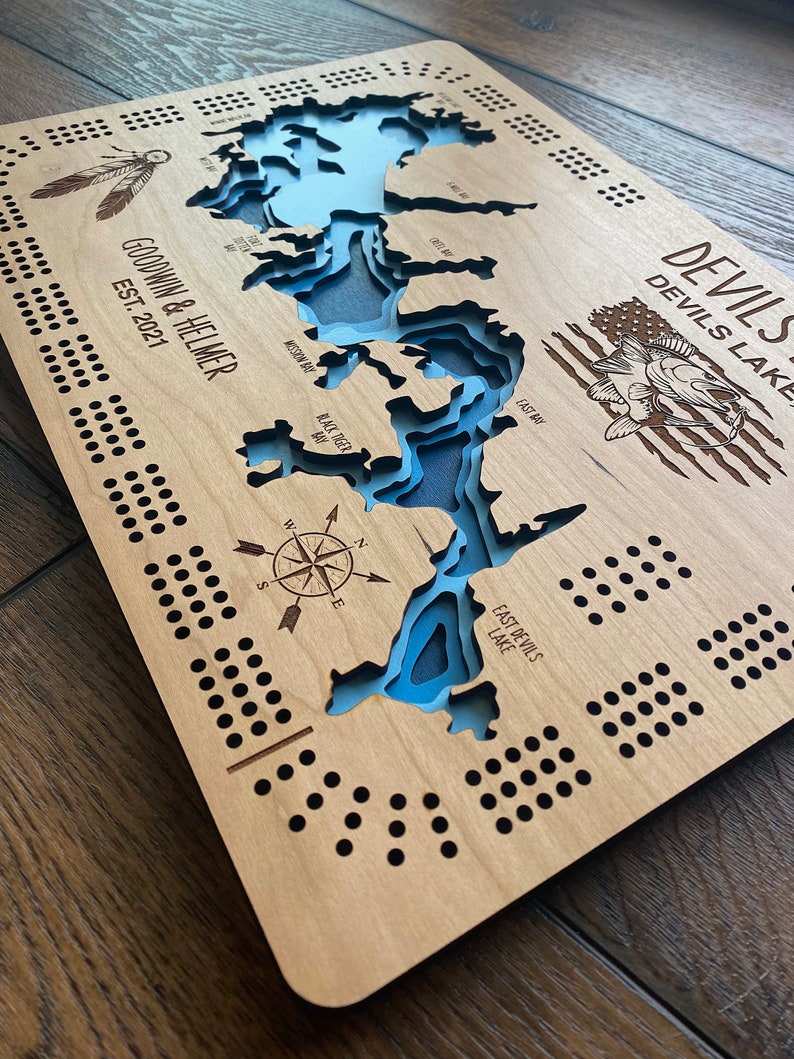 Pu&ograve; includere: Un tabellone di gioco in legno con una mappa del Devils Lake, che include una bussola, un pesce e il testo "GOODWIN & HELMER EST. 2021".