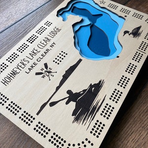 Pu&ograve; includere: Un tabellone da gioco in legno con un design di lago blu e il testo "Hohmeyer's Lake Clear Lodge Lake Clear, NY". Il tabellone ha una rosa dei venti e una silhouette di una persona in canoa.
