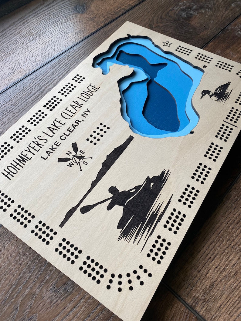 Pu&ograve; includere: Un tabellone da gioco in legno con un design di lago blu e il testo "Hohmeyer's Lake Clear Lodge Lake Clear, NY". Il tabellone ha una rosa dei venti e una silhouette di una persona in canoa.