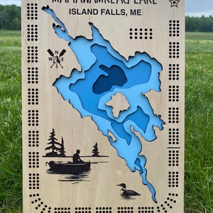 Pu&ograve; includere: Una mappa in legno del lago Mattawamkeag, Island Falls, ME. La mappa &egrave; blu e bianca e presenta una bussola, una barca con una persona che pesca e un tuffatore. La mappa &egrave; circondata da un bordo di piccoli punti.
