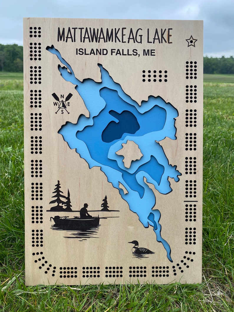 Pu&ograve; includere: Una mappa in legno del lago Mattawamkeag, Island Falls, ME. La mappa &egrave; blu e bianca e presenta una bussola, una barca con una persona che pesca e un tuffatore. La mappa &egrave; circondata da un bordo di piccoli punti.
