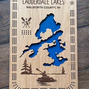 Pu&ograve; includere: Una mappa in legno di Lauderdale Lakes, contea di Walworth, WI. La mappa &egrave; blu e presenta una bussola, una stella e una silhouette di una persona in canoa. La mappa &egrave; circondata da un bordo di piccoli fori.