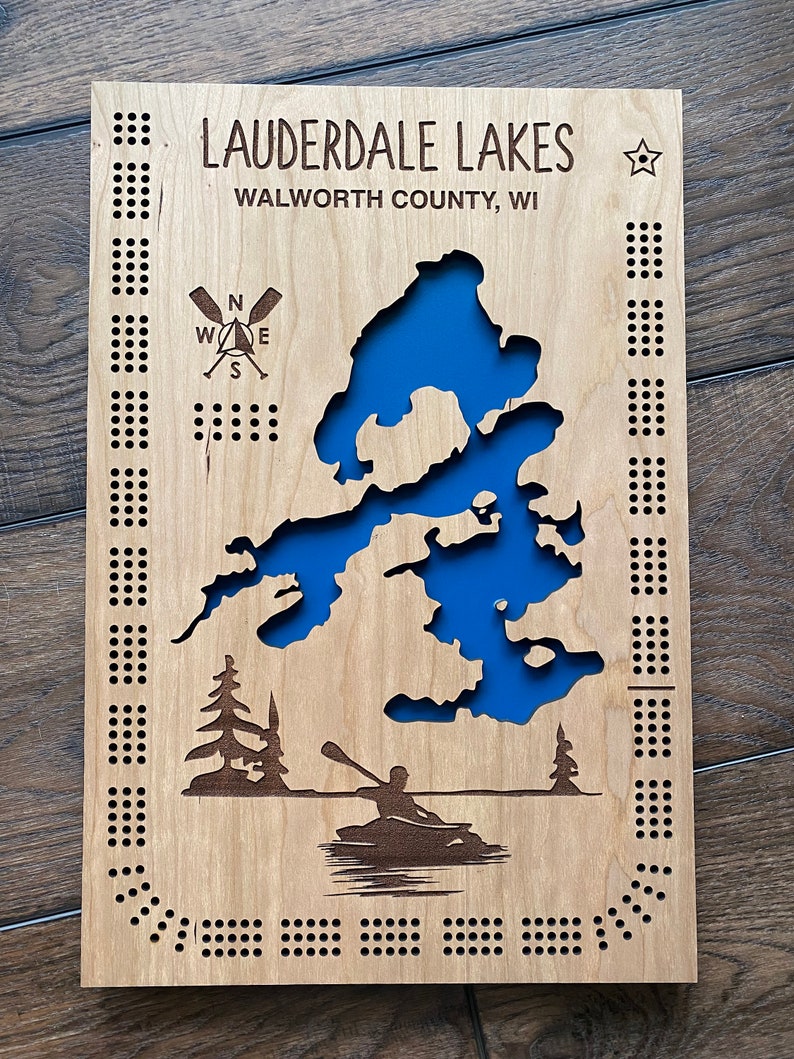 Pu&ograve; includere: Una mappa in legno di Lauderdale Lakes, contea di Walworth, WI. La mappa &egrave; blu e presenta una bussola, una stella e una silhouette di una persona in canoa. La mappa &egrave; circondata da un bordo di piccoli fori.
