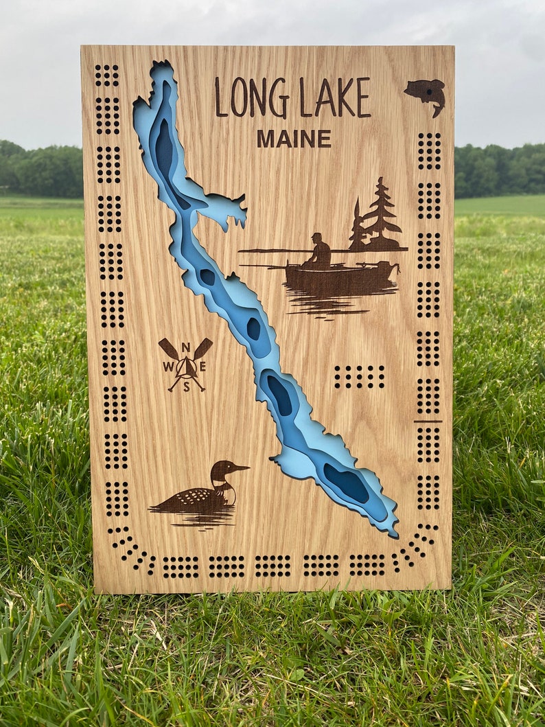 Pu&ograve; includere: Un tabellone di gioco in legno con una mappa del Long Lake, Maine. Il tabellone &egrave; dipinto di blu e ha una bussola, un loon e una scena di pesca. Il tabellone ha una griglia di buchi attorno al perimetro.
