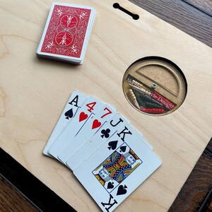 Pu&ograve; includere: Un tagliere di legno con un foro rotondo al centro. Il foro contiene un piccolo disco di legno con la scritta "Norffle Style Design" stampata su di esso. Ci sono cinque carte da gioco sul tagliere, tra cui un Asso di Picche, un 4 di Cuori, un 7 di Fiori, un Jack di Picche e un Re di Cuori. C'&egrave; anche un mazzo di carte nell'angolo in alto a sinistra del tagliere.
