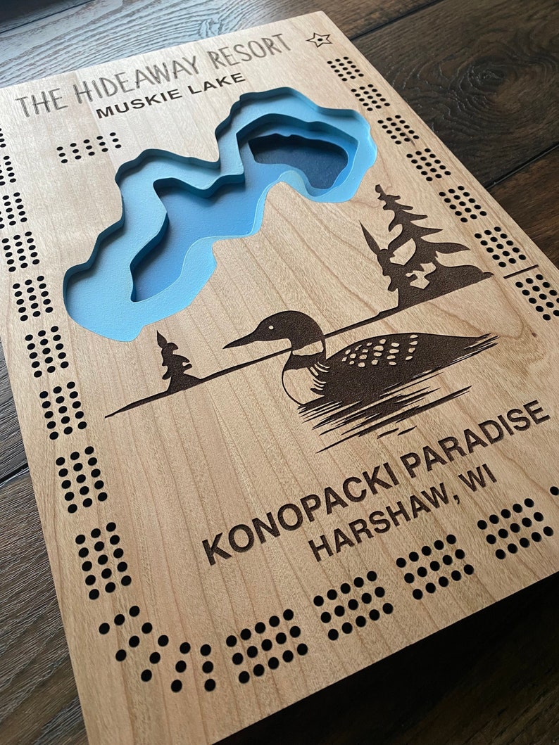 Pu&ograve; includere: Un tabellone da gioco in legno con un'incisione di un lago e un tuffatore. Il tabellone &egrave; decorato con punti neri e il testo "The Hideaway Resort Muskie Lake KonoPacki Paradise Harshaw, WI".