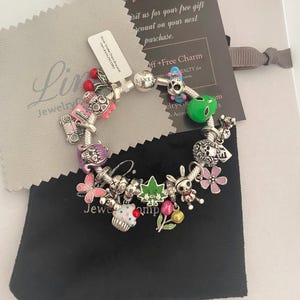 May include: A silver charm bracelet featuring a range of colourful charms, including a cupcake, a skull, a butterfly, a bunny, and a green alien. The bracelet is displayed on a black velvet pouch with the text "Linx Jewelry Company" on the tag.