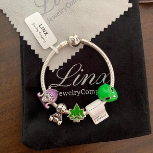May include: A silver charm bracelet featuring a selection of charms, including a skull, a marijuana leaf, a toilet roll with the text "Shit Happens", and an alien. The bracelet is displayed on a black velvet pouch with the text "Linx Jewelry Company" on it.