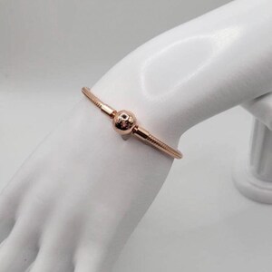 May include: A rose gold snake chain bracelet with a round charm. The bracelet is on a white mannequin hand.
