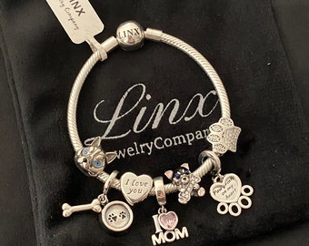 Puede incluir: Pulsera de plata con varios dijes con temática de perros, que incluyen huellas de patas, un hueso, una silueta de perro y un dije en forma de corazón que dice "Te amo". La pulsera también incluye un dije que dice "Huellas de patas en mi corazón" y un dije que dice "Yo Mamá".