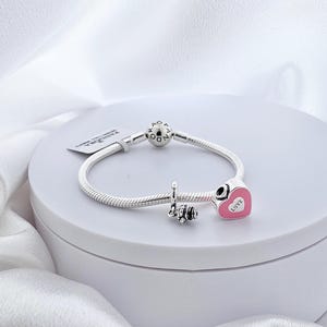 Könnte beinhalten: Ein silbernes Armband mit einem herzförmigen Anhänger und dem Wort "Love" in Weiß. Das Armband hat auch einen Anhänger mit dem Wort "Love" und einen kleinen Anhänger mit einem dekorativen Design. Das Armband wird auf einer weißen Oberfläche präsentiert.