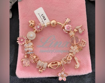 May include: A rose gold charm bracelet with a variety of charms, including flowers, a ladybug, a turtle, a heart, and an infinity symbol. The bracelet is on a pink velvet pouch with the text "LINX Jewelry Company".