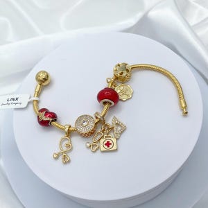 May include: Gold charm bracelet with red and gold charms, including a heart, a medical symbol, and a first aid kit. The bracelet is on a white background.