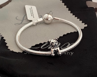Może przedstawiać: Srebrna bransoletka z zawieszką w kształcie psa. Bransoletka leży na czarnym aksamitnym woreczku z napisem "The Jewelry Company".
