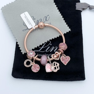 May include: Rose gold snake chain bracelet with pink charms, including hearts and beads. The bracelet is displayed on a black velvet pouch and a grey cleaning cloth. The tag reads "Rose Gold Snake Chain Bracelet".