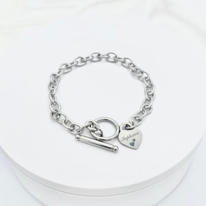 May include: A silver-colored chain bracelet with a toggle clasp. A heart-shaped charm is engraved with the name "Stephanie" and two small hearts. The bracelet is displayed on a white surface.