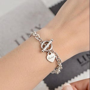 May include: A silver-toned chain link bracelet featuring a toggle clasp closure and a small heart-shaped charm. The charm is engraved with the name Racheal in a cursive script font. The bracelet is constructed from interconnected circular links.
