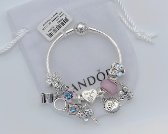 May include: A silver bracelet with multiple charms, including a flower, a key, a heart with the word "wife", and a pink bead. The bracelet is on a white drawstring bag.