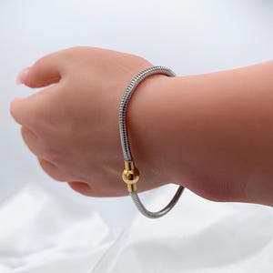 May include: A silver-toned bracelet with a flexible, ribbed design. The bracelet features a gold-colored clasp. The jewelry is shown on a person's wrist.