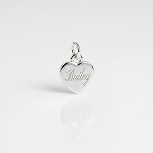 May include: A polished silver heart-shaped charm featuring the name Bailey engraved in a cursive script font. The charm includes a small circular jump ring at the top for attachment, reflecting a bright metallic finish on its smooth surface.