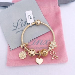 May include: A gold bracelet with charms, including a heart, a flower, a bee, and a key. The bracelet is on a pink velvet pouch with the text "Linx Jewelry Company" on a white tag.