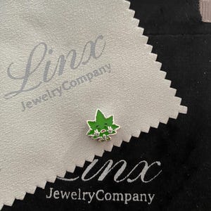 Op de afbeelding: Een kleine groene marihuana-bladbedel met een cartoon gezicht en opgeheven armen in een overgavepose. De bedel ligt op een witte juwelenpoetsdoek met de tekst "Linx Jewelry Company" erop gedrukt.