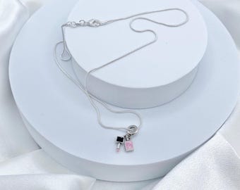 May include: A silver-colored necklace with a delicate chain. The pendant features two small charms: a black square and a pink square with a small pink lipstick charm. The necklace is displayed on a white, layered surface.