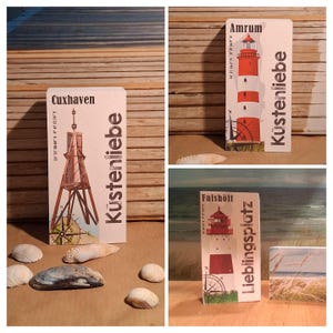 Könnte beinhalten: Drei rechteckige Postkarten mit Küstenmotiven. Eine zeigt eine Holzkonstruktion in Cuxhaven, eine weitere einen rot-weißen Leuchtturm in Amrum und die dritte einen Leuchtturm in Falshöft. Jede Karte trägt das Wort "Küstenliebe" und eine Windrose.
