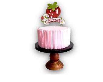 May include: A pink cake with a strawberry and the number 1 on top. The cake is on a black cake stand with a wooden base. The cake is decorated with a banner that says "Bethany".
