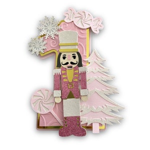 May include: A pink and gold papercraft number one with a nutcracker, a candy cane, and a snowflake design.