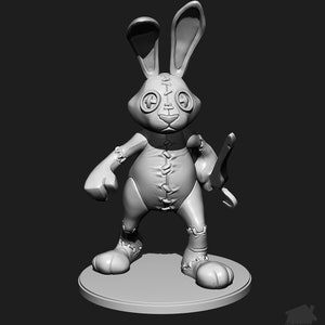 Op de afbeelding: Een grijs, gestikt, konijnvormig speelgoed met grote ogen en een scheve glimlach. Het speelgoed houdt een klein, houten, katapult in zijn rechterhand. Het speelgoed staat op een ronde, grijze basis.