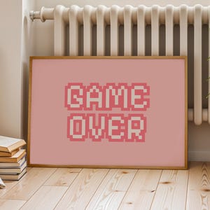 Könnte beinhalten: Ein gerahmter Druck mit den Worten "GAME OVER" in einer pixeligen Schriftart. Der Text ist in Rosa- und Weißtönen auf einem rosa Hintergrund gehalten. Der Rahmen ist hellbraun. Der Druck befindet sich auf einem Holzboden.