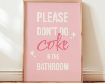 May include: A framed pink poster with the text "PLEASE DON'T DO coke IN THE BATHROOM" in white and pink lettering. The poster is in a light wood frame and is leaning against a white wall on a wooden floor.