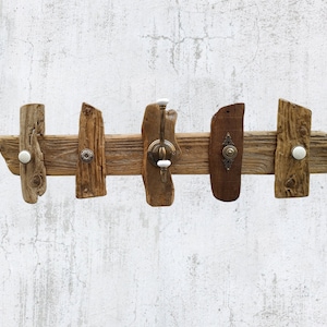 Op de afbeelding: Een rustieke houten kapstok met vijf haken. De kapstok is gemaakt van verweerde stukken drijfhout en heeft decoratieve knoppen en haken in verschillende stijlen, waaronder witte keramiek en antiek messing. De houttinten variëren van licht- tot donkerbruin.