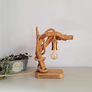 Op de afbeelding: Een rustieke houten tafellamp met een ontwerp van drijfhout. De lamp heeft een gloeilamp die aan een messing fitting hangt, met touwdetails. De basis is een rechthoekig houten blok. Een uniek woondecoratie-item, ongeveer 30 cm hoog.