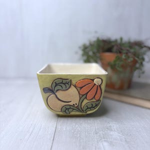 May include: A square, ceramic bowl with a light green exterior and a cream-colored interior. The bowl features a raised design of a peach and an orange flower with green leaves. The bowl is likely for decorative or serving purposes.