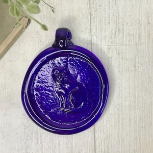 May include: A deep blue glass pendant with a cat silhouette. The pendant is round with a textured surface and a loop at the top for hanging. The cat is sitting, with its tail curled.