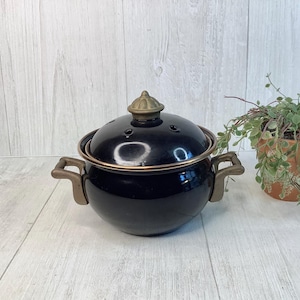 May include: A black enamel cooking pot with a lid and two handles. The pot has a gold-colored rim and handles. The lid has a decorative knob and several small holes. A small potted plant is in the background.