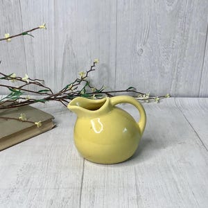 May include: A small, round, yellow ceramic pitcher with a handle and a spout. The pitcher is sitting on a light-colored wooden surface. A book and sprigs of flowers are in the background.
