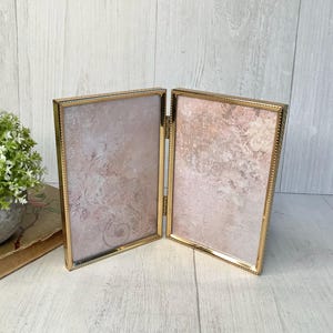 May include: A vintage-style, double-hinged photo frame with a gold-colored metal frame. The frame holds two rectangular inserts with a soft, pink and beige floral design. The frame is displayed on a white wooden surface.