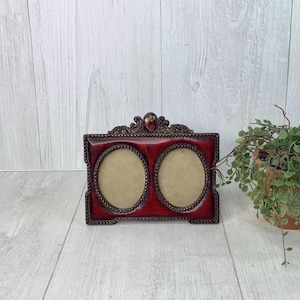 May include: A decorative, rectangular photo frame with two oval openings. The frame is a deep red with ornate gold-colored trim and a decorative top. The frame is designed to hold two photos and is a vintage style.