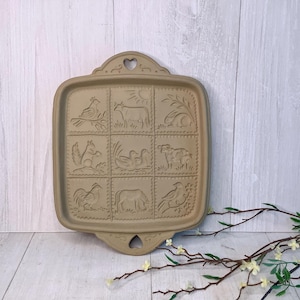 May include: Tan ceramic baking mold with embossed farm animal designs. The square mold features a rooster, cow, squirrel, ducks, sheep, horse, and bird. The top has a heart-shaped cutout.