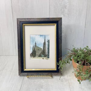 May include: Framed watercolor print of a cathedral with tall spires and architectural details. The artwork is set within a black and gold frame, displayed on a decorative stand. A potted plant with green foliage is in the background.
