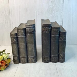 May include: Two sets of three dark gray book-shaped decorative objects featuring embossed floral patterns on the spines. Each set consists of three vertical rectangular blocks arranged side by side, mimicking the appearance of antique leather-bound books.