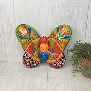 May include: A colorful ceramic butterfly with a vibrant design. The wings are green, orange, and blue, with a black and white pattern on the lower sections. The butterfly is a decorative item.