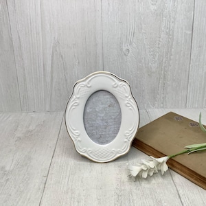 May include: An oval-shaped white picture frame with a gold-toned rim and embossed scrollwork details. The frame features a central opening with a gray patterned insert, and the outer edges have a scalloped design with a textured grid pattern at the base.