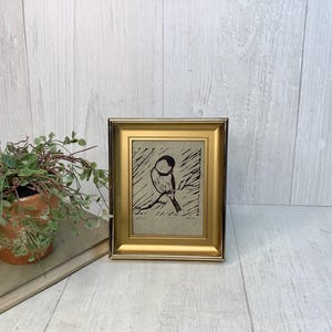 May include: A framed art print featuring a black ink linocut illustration of a small bird perched on a branch. The artwork is set within a metallic gold frame with a beveled edge, showcasing a minimalist aesthetic on a light-colored paper background.