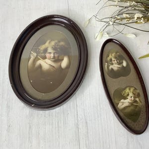 May include: Two antique oval-shaped picture frames. One frame is a dark brown color and contains a sepia-toned image of a cherub holding a bow and arrow. The other frame is a long, narrow shape with two cherub images.